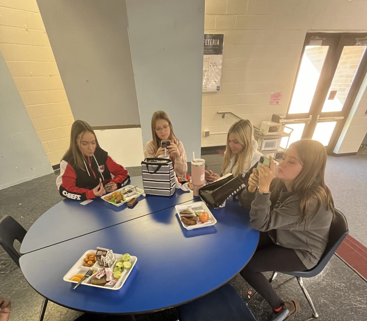 Kinley Martinek. Blakley Walter, Averi Broxterman, and Giuliana Dawkins, sit at a table during lunch, all disconnected with each other on their phones.